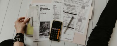 Top view of desk with papers, calculator, and pen, with an arm and leg partially in frame